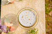top down image of lavendar sweet heather anne cake on class stand with flowers in the background during styled shoot