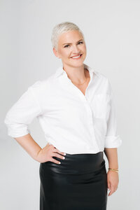 Amy Dewaele, a real estate agent, in a white shirt and black skirt, smiling against a light blue background.