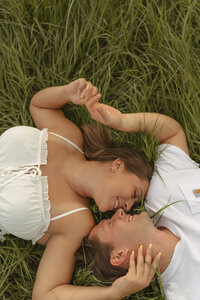 Couple laying in tall green grass, smiling and gazing at each other during a candid and intimate moment.