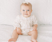 one year old little girl in cream colored dress sitting on a bed with a bow in her hair smiling 
