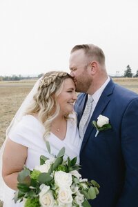 A groom kisses a bride's forehead as she turns towards him holding her bouquet.