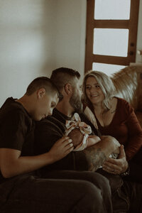 A family of 4 sit on their living room couch welcoming their newest addition. Big brother is lightly stroking baby's head while mom and dad look into each others eyes, a soft smile on their lips.