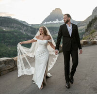 Bride and groom walking hand in hand along Going-to-the-Sun Road in Glacier National Park, with mountains in the background and the bride’s veil flowing in the wind.