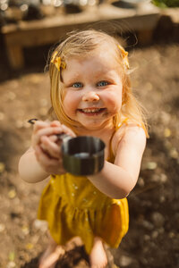 toddler girl holds measuring cup up as she smiles at the camera while playing in a mud kitchen