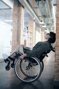 A woman wearing a blazer, khakis, and glasses sits in a wheelchair, leaning back against a brick wall. She has a laptop in her lap, head tilted up with eyes closed.