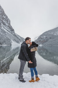Newly engaged couple sharing a kiss with reflections of Lake Louise in the water.