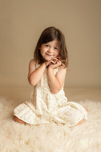 Little girl wearing a cream colored dress smiles as she snuggles a small stuffed animal during her portrait session.