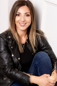 Company branding portrait Three Stories Jewelry Owner Camelia Chiriac sitting against white wall hands around knee while smiling wearing denim with black leather jacket