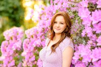 Red-headed Charlotte Area Senior stands surrounded by lilac blooms in a purple dress with the sun glowing behind her during her graduate photo session