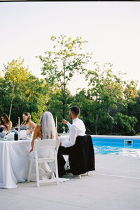 poolside wedding reception, Documentary wedding photography, bride in white dress, groom and bridesmaid sitting at table next to blue in-ground pool