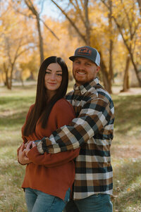 A man holds his wife from behind as they both smile at the camera. They're in a park in Idaho Falls, Idaho.