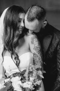 A black and white photo from a San Francisco photoshoot of a bride and groom sharing a tender moment. The groom, with tattoos on his arms, gently kisses the bride on her shoulder.