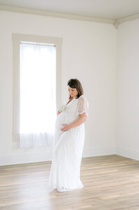 A maternity client poses in a natural light studio wearing a white dress make of delicate lace.
