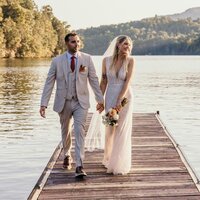 a newlywed couple holding hands and walking on a dock on a lake 