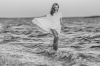 Woman with long, wavy, blonde hair walks in a short white dress along the shallow ocean shoreline.