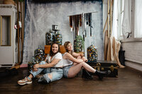 Portrait of two women sitting back-to-back during a film lab branding session. Surrounded by large jars filled with used film canisters, a vintage film camera, and hanging film strips, the creative workspace exudes a vibrant, artistic energy. The women, dressed casually in white tops and denim, are smiling and relaxed, showcasing the welcoming and innovative atmosphere of the film lab. The natural lighting and textured backdrop emphasize the authenticity and passion behind their craft.