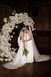 Two brides in elegant white wedding gowns sharing a kiss beneath a stunning floral arch during their intimate and romantic indoor wedding ceremony, capturing love and celebration