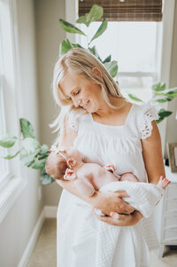 Mother in white dress cradles  newborn baby girl during in-home photography session