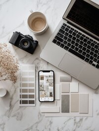 Flat lay of creative workspace on marble desk with open laptop, black camera, cup of coffee, neutral color-swatch cards, tile samples and smartphone displaying mood board images