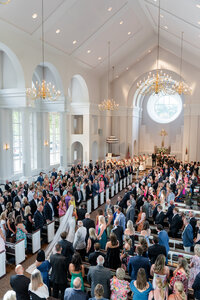A bride and her father walking down the aisle during a church ceremony. With large windows letting light shine through.
