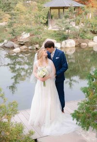 A bride and groom in a blue suit smile at each other near a lake in the summertime at Fredrick Meijer Gardens in Grand Rapids.