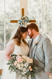 A bride and groom stand forehead to forehead during their indoor wedding ceremony at Ashton Gardens North in Houston, Texas. The couple is framed by floral arrangements and a wooden cross in the background, creating a romantic and intimate moment.