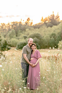 Pregnant Woman standing next to man. They are cheek-to-cheek and both holding baby bump during Oregon Maternity Photography session outdoors.