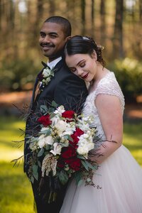 A bride holds her red and white floral bouquet by Flowers  and leans against the groom.