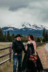 Bride in a black dress and groom in a cowboy hat standing hand in hand on a gravel path with Mount Shasta rising behind them under stormy skies.