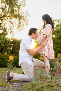 Romantic golden hour photo of dad kneeling to kiss his partner's baby bump in a garden, captured by Ellobelle Photography Family Photographer