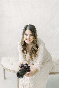 Photographer Ana Dufreche wears an ivory dress while holding a professional camera, smiling in front of a cream background.