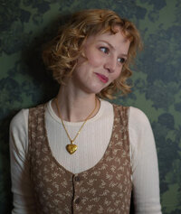 A person with short, curly blonde hair and wearing a white long-sleeve shirt under a brown, floral-patterned vest stands in front of a dark green, foliage-inspired wallpaper. They have a gold heart-shaped pendant necklace and are glancing off to the side while smiling, much like one would after perfecting their Showit template customization.