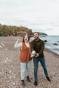 A couple standing on the beach laughing and popping a bottle of champagne 