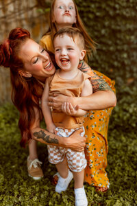 mom wearing a yellow floral dress embraces son from behind as they giggle at each other in front  of ivy wall at local coffee shop