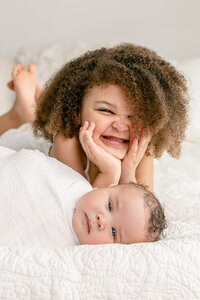Toddler girl in floral dress sitting crossed-legged and holding swaddled baby brother in her arms. Baby is sleeping and she is bending over him to kiss him on the side of the face. Sweet, tender moment between new siblings.