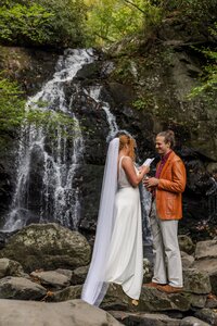 A bride and groom stand in front of a large, tumbling waterfalls reading their vows to each other. The bride is in a flowy wedding dress with a long veil and the groom is wearing tan pants with an orange leather jacket. They are surrounded by rocks and lush green trees.