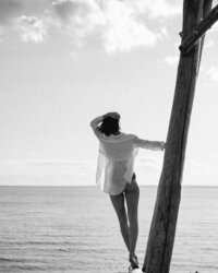 A woman in swimwear gazes out at the ocean, standing near a wooden post, evoking a sense of contemplation and connection with nature.
