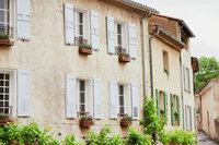 A charming, rustic building with light beige walls and white shutters. The windows have small balconies with potted plants, and there’s greenery along the bottom of the building, enhancing the quaint, picturesque ambiance.