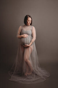 35 week pregnant woman dressed in gray sheer maternity dress on gray backdrop posing standing up holding her baby bump and looking to the side