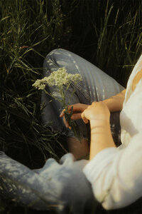 A woman sits in the grass, gently holding a flower and enjoying a peaceful moment outdoors.