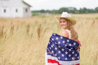 senior girl standing in wheat field turned looking back towards camera with flag wrapped around her with cowgirl hat on photographed by Jamie Lynette Photography canton senior photographer