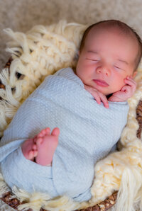 Newborn baby peacefully sleeping, wrapped in a light blue blanket and resting in a cozy basket with soft cream textures – captured by Capture Joy Photography, Northern Virginia family photographer