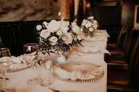 A sophisticated wedding table setting with white floral centerpieces, glass chargers with beaded rims, and knotted beige napkins on a crisp white tablecloth.