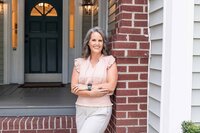 Business owner in a light peach shirt leanig up against a brick wall in front of her office building