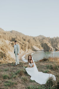 Bride crouched, groom behind looking at her on the edge of a cliff
