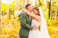 groom in green suit kissing bride's forehead  in wedding dress with veil, wedding bouquet over his back, in fall aspen grove in Flagstaff Arizona elopement