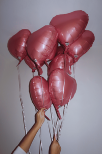 A person holding a bunch of pink heart-shaped balloons against a light background. The balloons are tied together with ribbons.
