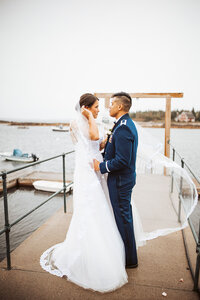 Bride and groom standing on a pier with flowing veil and boats in the background during their Maine coastal wedding.
