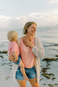 A mom wearing a pink tee shirt and a loose knit sweater holds her blonde toddler in a pink tee shirt on her hip while on the beach and laughing. 