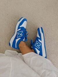 Person wearing bright blue and white New Balance sneakers on a neutral carpet, styled with a gold anklet and white pants. modern, casual lifestyle aesthetic.
